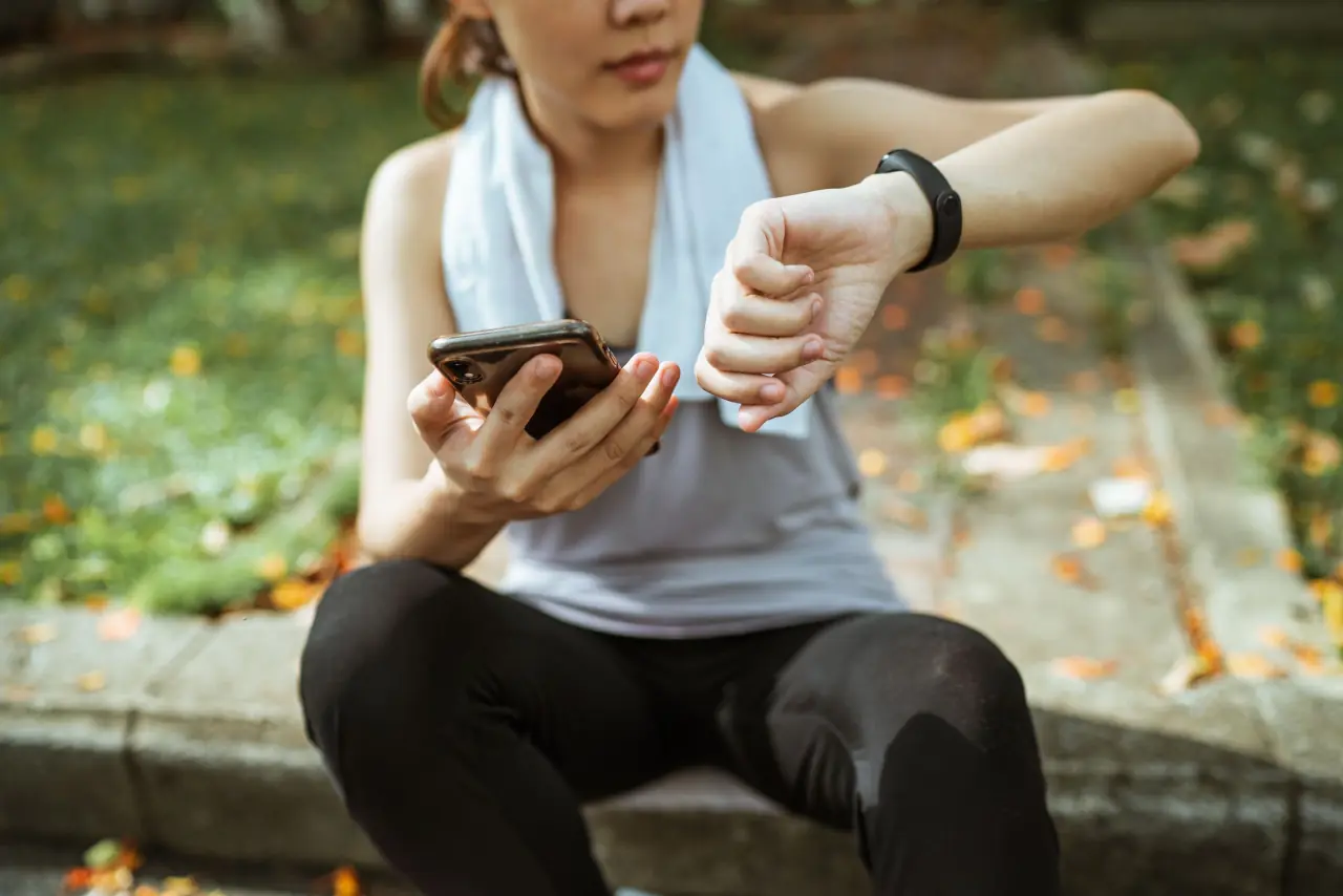 An athlete with a towel around her neck rests with a phone in one hand and checks her watch.