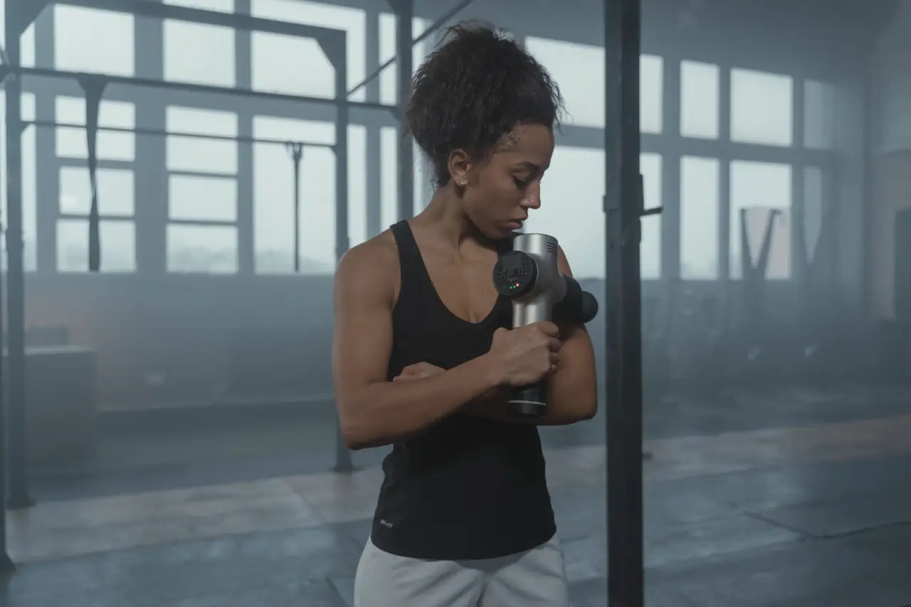 A woman works a massage gun over her forearm in a hazy, window-lit training hall.